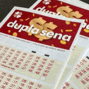 Foto em close de vários volantes de aposta da loteria "Dupla Sena", da Caixa Econômica Federal, sobrepostos. Os bilhetes têm o topo vermelho com o nome do jogo escrito em branco e um cifrão estilizado em dourado ao fundo, cercado por pequenas moedas. Abaixo, há campos de preenchimento com tabelas numéricas organizadas em linhas e colunas, indo do número 01 ao 50.