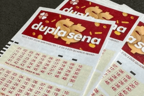Foto em close de vários volantes de aposta da loteria "Dupla Sena", da Caixa Econômica Federal, sobrepostos. Os bilhetes têm o topo vermelho com o nome do jogo escrito em branco e um cifrão estilizado em dourado ao fundo, cercado por pequenas moedas. Abaixo, há campos de preenchimento com tabelas numéricas organizadas em linhas e colunas, indo do número 01 ao 50.