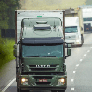 Fotografia em plano médio de um caminhão verde da marca Iveco trafegando em uma rodovia asfaltada. O caminhão está em primeiro plano, centralizado, com os faróis acesos e a placa visível na parte inferior. Ao fundo, em desfoque, aparecem outros dois caminhões grandes e um carro de passeio prata, todos seguindo na mesma direção da estrada, que é cercada por vegetação nas laterais em um dia nublado.