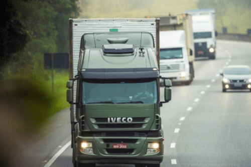 Fotografia em plano médio de um caminhão verde da marca Iveco trafegando em uma rodovia asfaltada. O caminhão está em primeiro plano, centralizado, com os faróis acesos e a placa visível na parte inferior. Ao fundo, em desfoque, aparecem outros dois caminhões grandes e um carro de passeio prata, todos seguindo na mesma direção da estrada, que é cercada por vegetação nas laterais em um dia nublado.