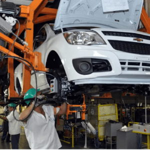 Fotografia do interior de uma fábrica de automóveis mostrando uma linha de montagem. Um carro branco da marca Chevrolet está suspenso por uma grande estrutura metálica laranja. O capô do veículo está aberto. Em primeiro plano, um operário usando capacete verde de proteção e luvas trabalha na parte inferior dianteira do carro. Ao fundo, outro operário com capacete semelhante opera maquinários na linha de produção.