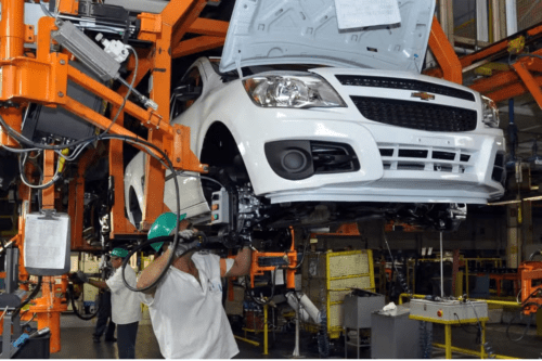Fotografia do interior de uma fábrica de automóveis mostrando uma linha de montagem. Um carro branco da marca Chevrolet está suspenso por uma grande estrutura metálica laranja. O capô do veículo está aberto. Em primeiro plano, um operário usando capacete verde de proteção e luvas trabalha na parte inferior dianteira do carro. Ao fundo, outro operário com capacete semelhante opera maquinários na linha de produção.