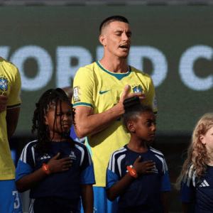 Dois jogadores da seleção brasileira de futebol masculino e três crianças que os acompanham estão perfilados em um campo, de frente, com a mão direita sobre o peito, cantando o hino nacional. Os jogadores vestem a camisa amarela tradicional do Brasil e calções azuis. As crianças à frente vestem camisetas azuis escuras com detalhes em branco. Ao fundo, um painel digital exibe parcialmente as palavras "WORLD CUP" em letras maiúsculas brancas.