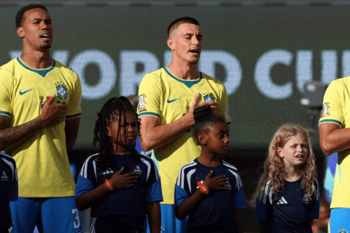 Dois jogadores da seleção brasileira de futebol masculino e três crianças que os acompanham estão perfilados em um campo, de frente, com a mão direita sobre o peito, cantando o hino nacional. Os jogadores vestem a camisa amarela tradicional do Brasil e calções azuis. As crianças à frente vestem camisetas azuis escuras com detalhes em branco. Ao fundo, um painel digital exibe parcialmente as palavras "WORLD CUP" em letras maiúsculas brancas.