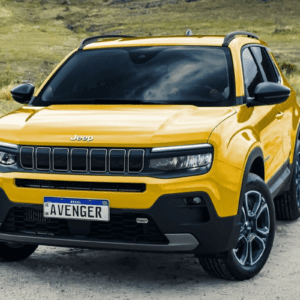 Uma fotografia em ângulo de três quartos de um carro SUV compacto Jeep Avenger amarelo vibrante. O veículo está estacionado em um terreno de terra batida e areia, com uma colina gramada ao fundo. O SUV tem o teto, as colunas, os espelhos retrovisores e os para-choques pretos. Na frente, destaca-se a icônica grade de sete fendas da Jeep e uma placa de identificação no padrão Mercosul (Brasil) escrita "AVENGER".