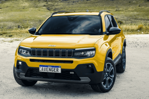 Uma fotografia em ângulo de três quartos de um carro SUV compacto Jeep Avenger amarelo vibrante. O veículo está estacionado em um terreno de terra batida e areia, com uma colina gramada ao fundo. O SUV tem o teto, as colunas, os espelhos retrovisores e os para-choques pretos. Na frente, destaca-se a icônica grade de sete fendas da Jeep e uma placa de identificação no padrão Mercosul (Brasil) escrita "AVENGER".