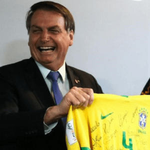 O ex-presidente Jair Bolsonaro aparece sorrindo e rindo abertamente enquanto segura com as duas mãos uma camisa amarela oficial da Seleção Brasileira de Futebol. Ele veste um terno escuro, camisa social clara e gravata azul. A camisa de futebol, que está autografada por vários jogadores, exibe o escudo da CBF e o número 4 em verde. Ao fundo, uma parede clara e, à direita, parte de uma camisa preta com o logotipo da marca Itaú.