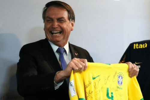 O ex-presidente Jair Bolsonaro aparece sorrindo e rindo abertamente enquanto segura com as duas mãos uma camisa amarela oficial da Seleção Brasileira de Futebol. Ele veste um terno escuro, camisa social clara e gravata azul. A camisa de futebol, que está autografada por vários jogadores, exibe o escudo da CBF e o número 4 em verde. Ao fundo, uma parede clara e, à direita, parte de uma camisa preta com o logotipo da marca Itaú.