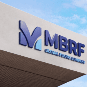 Foto em ângulo contra-plongé do letreiro de uma empresa em uma fachada moderna de concreto claro, sob um céu azul com nuvens esparsas. À esquerda, há um logotipo estilizado formado pela letra "M" azul-escura com um detalhe em azul-claro na base. À direita, constam as siglas "MBRF" em letras grandes e azuis, e logo abaixo a inscrição "GLOBAL FOODS COMPANY" em letras menores, também em azul.