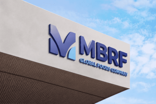 Foto em ângulo contra-plongé do letreiro de uma empresa em uma fachada moderna de concreto claro, sob um céu azul com nuvens esparsas. À esquerda, há um logotipo estilizado formado pela letra "M" azul-escura com um detalhe em azul-claro na base. À direita, constam as siglas "MBRF" em letras grandes e azuis, e logo abaixo a inscrição "GLOBAL FOODS COMPANY" em letras menores, também em azul.