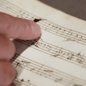 Uma pessoa aponta com o dedo indicador para uma mancha escura de tinta em uma partitura musical antiga, escrita à mão.