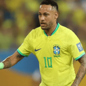 Fotografia em plano médio do jogador de futebol Neymar em campo, vestindo a camiseta amarela oficial da Seleção Brasileira com o número 10 verde no peito. Ele está focado, olhando para o lado esquerdo da imagem, com o braço direito estendido e uma faixa verde clara no pulso. O fundo mostra as arquibancadas de um estádio desfocadas.