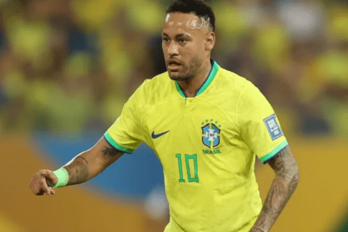 Fotografia em plano médio do jogador de futebol Neymar em campo, vestindo a camiseta amarela oficial da Seleção Brasileira com o número 10 verde no peito. Ele está focado, olhando para o lado esquerdo da imagem, com o braço direito estendido e uma faixa verde clara no pulso. O fundo mostra as arquibancadas de um estádio desfocadas.