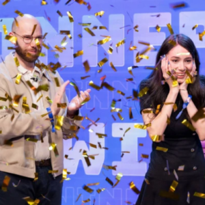 Uma foto de plano médio captura um momento de celebração em um palco, sob uma chuva de confetes dourados brilhantes. À esquerda, um homem calvo com óculos, barba e jaqueta bege aplaude sorrindo. À direita, uma mulher de cabelos longos e escuros, vestindo um vestido preto, expressa surpresa e alegria genuínas, segurando o rosto com as duas mãos e sorrindo amplamente. O fundo é composto por um painel azul vibrante com a palavra "WINNER" (Vencedor) repetida em letras grandes e estilizadas.