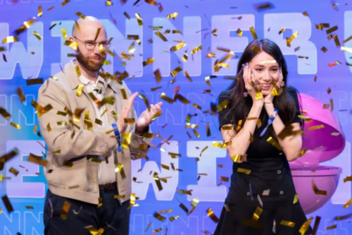 Uma foto de plano médio captura um momento de celebração em um palco, sob uma chuva de confetes dourados brilhantes. À esquerda, um homem calvo com óculos, barba e jaqueta bege aplaude sorrindo. À direita, uma mulher de cabelos longos e escuros, vestindo um vestido preto, expressa surpresa e alegria genuínas, segurando o rosto com as duas mãos e sorrindo amplamente. O fundo é composto por um painel azul vibrante com a palavra "WINNER" (Vencedor) repetida em letras grandes e estilizadas.