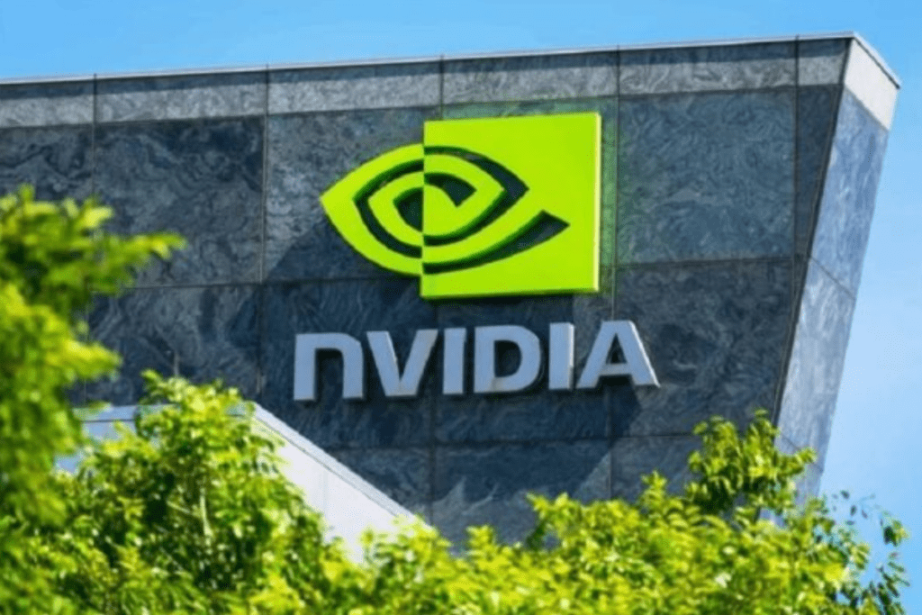 Uma imagem em plano médio mostra a fachada do edifício da sede da NVIDIA Corporation sob um céu azul claro. O edifício apresenta uma parede texturizada de pedra cinza-escura com grandes janelas espelhadas. Destacado proeminentemente na parede está o logotipo da NVIDIA: um símbolo verde-limão em formato de olho espiralado contido em um quadrado, posicionado acima do nome da marca "NVIDIA" em letras tridimensionais prateadas. Na parte inferior da imagem, folhas verdes vibrantes de árvores obscurecem parcialmente a base do edifício.