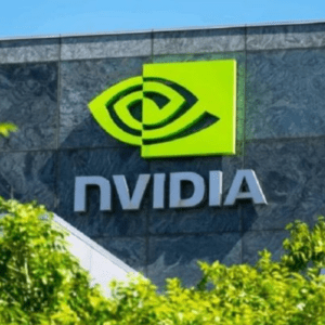 Uma imagem em plano médio mostra a fachada do edifício da sede da NVIDIA Corporation sob um céu azul claro. O edifício apresenta uma parede texturizada de pedra cinza-escura com grandes janelas espelhadas. Destacado proeminentemente na parede está o logotipo da NVIDIA: um símbolo verde-limão em formato de olho espiralado contido em um quadrado, posicionado acima do nome da marca "NVIDIA" em letras tridimensionais prateadas. Na parte inferior da imagem, folhas verdes vibrantes de árvores obscurecem parcialmente a base do edifício.