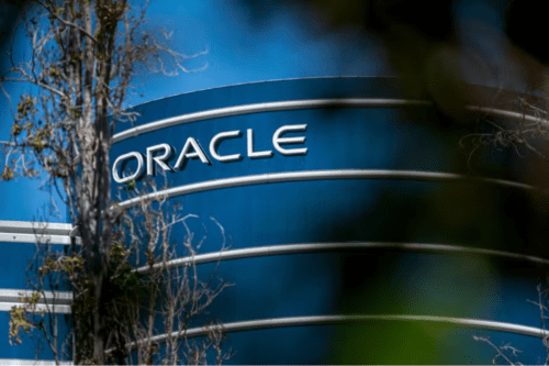 Foto em ângulo contra-plongée mostrando o topo de um edifício comercial cilíndrico de vidro azul com o logotipo da empresa "ORACLE" em letras maiúsculas brancas. Galhos de árvores desfocados aparecem em primeiro plano nas laterais.