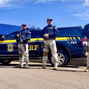 Três policiais da Polícia Rodoviária Federal (PRF) do Brasil posam uniformizados em frente a uma viatura preta e amarela. Os policiais vestem gandolas escuras, coletes balísticos, calças táticas na cor caqui e botas. Dois deles seguram armas longas táticas. A viatura exibe o brasão da PRF na porta e o número de emergência 191 na lateral traseira. O cenário é ao ar livre, sob um céu azul com poucas nuvens.