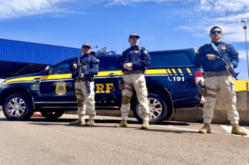 Três policiais da Polícia Rodoviária Federal (PRF) do Brasil posam uniformizados em frente a uma viatura preta e amarela. Os policiais vestem gandolas escuras, coletes balísticos, calças táticas na cor caqui e botas. Dois deles seguram armas longas táticas. A viatura exibe o brasão da PRF na porta e o número de emergência 191 na lateral traseira. O cenário é ao ar livre, sob um céu azul com poucas nuvens.