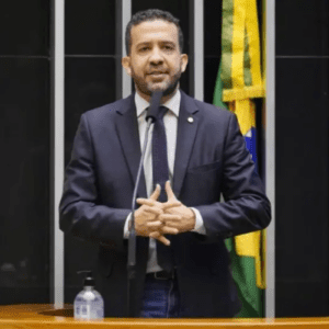 Um homem de terno escuro, camisa branca e gravata fala ao microfone em um púlpito de madeira na Câmara dos Deputados. Ele tem cabelos curtos e barba aparada, e está com as mãos entrelaçadas à frente do corpo enquanto discursa. Ao fundo, há uma parede com faixas verticais escuras e claras, além de parte da bandeira do Brasil visível à direita. Sobre a bancada, vê-se um frasco de álcool em gel e um copo de vidro.