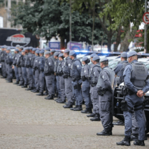 Uma longa fileira de policiais militares da Força Tática perfilados em uma praça pública, ao lado de viaturas brancas com girofles ligados.