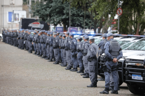 Uma longa fileira de policiais militares da Força Tática perfilados em uma praça pública, ao lado de viaturas brancas com girofles ligados.