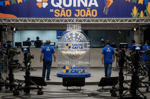 Fotografia em plano médio do estúdio de gravação do sorteio da loteria "Quina de São João", promovida pelas Loterias Caixa. No centro, destaca-se um grande globo acrílico transparente cheio de bolinhas numeradas. Dois homens de costas, vestindo camisetas azuis com o logotipo das Loterias Caixa e calças jeans, estão posicionados nas laterais do globo. Na parte superior, há um grande banner azul e amarelo com a inscrição "QUINA DE SÃO JOÃO", decorado com elementos festivos juninos, como bandeirinhas e espigas de milho. O primeiro plano exibe câmeras de transmissão profissional em tripés e cabos espalhados pelo chão, enquanto ao fundo observa-se a sala de controle com monitores e técnicos trabalhando.