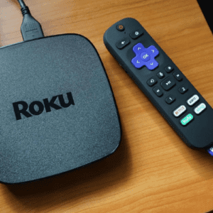 Dispositivo de streaming Roku na cor grafite com cabo conectado e controle remoto Roku ao lado, sobre superfície de madeira.