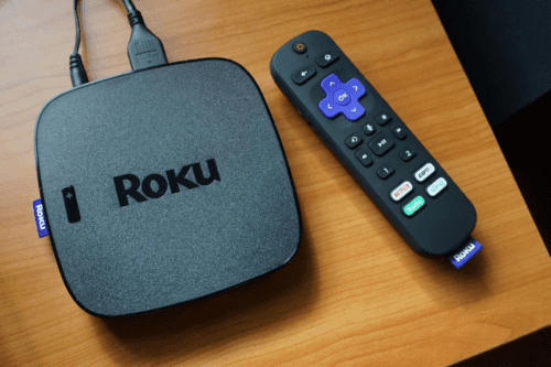 Dispositivo de streaming Roku na cor grafite com cabo conectado e controle remoto Roku ao lado, sobre superfície de madeira.