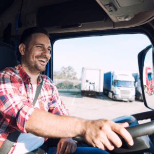 Fotografia em ângulo médio do interior da cabine de um caminhão. Um motorista homem, sorridente e de barba curta, veste camisa xadrez vermelha e branca e usa cinto de segurança enquanto segura o volante. Através da janela aberta do veículo, vê-se um pátio ensolarado com outros caminhões estacionados ao fundo.