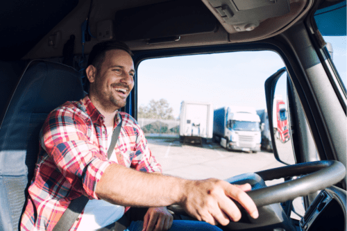 Fotografia em ângulo médio do interior da cabine de um caminhão. Um motorista homem, sorridente e de barba curta, veste camisa xadrez vermelha e branca e usa cinto de segurança enquanto segura o volante. Através da janela aberta do veículo, vê-se um pátio ensolarado com outros caminhões estacionados ao fundo.