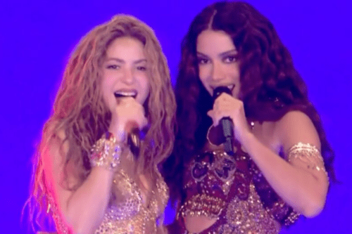Fotografia em plano médio das cantoras Shakira e Anitta dividindo o palco. À esquerda, Shakira sorri enquanto canta em um microfone, exibindo seus longos cabelos loiros e ondulados e um figurino dourado brilhante com pulseiras combinando. À direita, Anitta também segura um microfone próximo à boca, sorrindo sutilmente; ela tem cabelos longos, escuros e ondulados, e usa um figurino bordado em tons de vermelho e dourado com braceletes metálicos. O fundo da imagem é inteiramente azul-escuro.