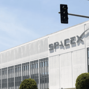 Fachada externa de um grande edifício branco com o letreiro cinza da empresa "SPACEX" em destaque. O prédio possui várias janelas retangulares organizadas em fileiras. Em primeiro plano, aparecem copas de árvores verdes decorativas e uma placa de trânsito à direita. Na parte superior, um semáforo preto está pendurado sob um céu azul claro com nuas poucas nuvens.