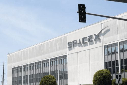 Fachada externa de um grande edifício branco com o letreiro cinza da empresa "SPACEX" em destaque. O prédio possui várias janelas retangulares organizadas em fileiras. Em primeiro plano, aparecem copas de árvores verdes decorativas e uma placa de trânsito à direita. Na parte superior, um semáforo preto está pendurado sob um céu azul claro com nuas poucas nuvens.