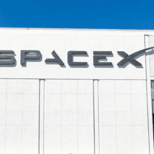 Fachada de um edifício branco com o logotipo estilizado da empresa "SPACEX" fixado em letras escuras no topo. O céu acima está limpo e azul. Nas laterais inferiores, são visíveis partes de janelas espelhadas e o topo de algumas árvores.