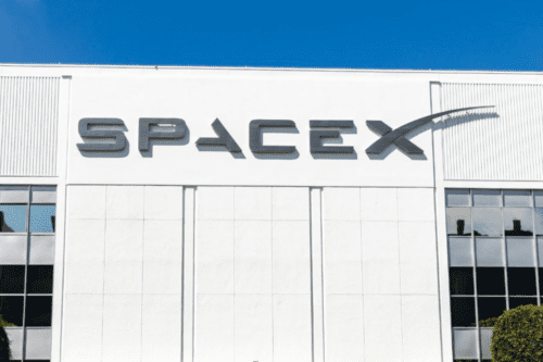 Fachada de um edifício branco com o logotipo estilizado da empresa "SPACEX" fixado em letras escuras no topo. O céu acima está limpo e azul. Nas laterais inferiores, são visíveis partes de janelas espelhadas e o topo de algumas árvores.