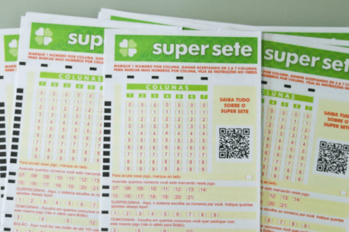 Três volantes oficiais de papel da loteria "Super Sete", da Caixa Econômica Federal, dispostos lado a lado sobre uma superfície clara. Os bilhetes possuem detalhes em verde e trazem a tabela principal dividida em 7 colunas numeradas de 1 a 7, com opções de números de 0 a 9 para marcar em cada uma delas. No canto superior esquerdo de cada volante, há o logotipo de um trevo de quatro folhas branco dentro de um quadrado verde, seguido pelo nome do jogo escrito em letras minúsculas brancas. Do lado direito da tabela de números, há um código QR em preto e branco.