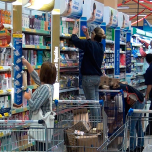 Interior de mercado com várias prateleiras de produtos de higiene e limpeza, pessoas escolhendo itens e carrinhos de compras com caixas e sacolas em destaque.