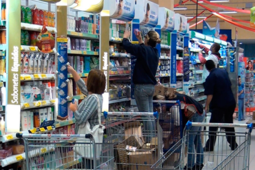 Interior de mercado com várias prateleiras de produtos de higiene e limpeza, pessoas escolhendo itens e carrinhos de compras com caixas e sacolas em destaque.