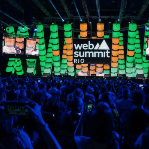 Uma grande plateia lota um auditório escuro durante o Web Summit Rio. Ao fundo, um palco iluminado exibe painéis de LED verticais em tons de verde e laranja. No centro do palco, um grande letreiro digital exibe o logotipo oficial do evento: "web summit RIO" em letras brancas. Telas suspensas nas laterais mostram os palestrantes sentados em poltronas brancas conversando. Em primeiro plano, várias pessoas na plateia seguram seus smartphones para registrar o evento.