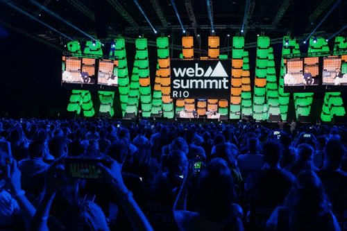 Uma grande plateia lota um auditório escuro durante o Web Summit Rio. Ao fundo, um palco iluminado exibe painéis de LED verticais em tons de verde e laranja. No centro do palco, um grande letreiro digital exibe o logotipo oficial do evento: "web summit RIO" em letras brancas. Telas suspensas nas laterais mostram os palestrantes sentados em poltronas brancas conversando. Em primeiro plano, várias pessoas na plateia seguram seus smartphones para registrar o evento.