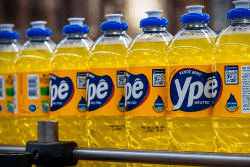Uma fileira de garrafas de detergente líquido da marca Ypê, do tipo "Neutro", alinhadas em uma esteira de produção industrial. O detergente tem uma cor amarela vibrante e transparente. As garrafas são de plástico transparente com rótulos amarelos e azuis, apresentando o logotipo da marca e tampas plásticas azuis com bico dosador branco. O foco está na garrafa mais à direita, com as outras ficando progressivamente desfocadas ao fundo.