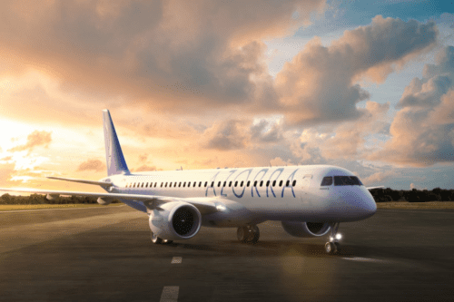 Uma aeronave comercial de passageiros branca com a inscrição "AZORRA" em letras azuis estilizadas na lateral da fuselagem. O avião está estacionado em uma pista de pouso asfaltada, de frente para o lado direito, com os faróis dianteiros acesos. O fundo mostra uma paisagem de vegetação baixa sob um céu dramático de fim de tarde ou amanhecer, com nuvens volumosas iluminadas por tons dourados, laranjas e rosados do sol.