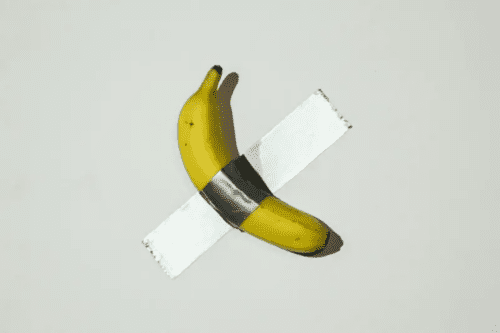 Uma banana amarela e madura colada diagonalmente em uma parede branca com um pedaço de fita adesiva prateada (duck tape). A obra está iluminada de forma a projetar uma sombra sutil na parede. Esta é a famosa obra de arte conceitual intitulada "Comedian", criada pelo artista italiano Maurizio Cattelan.
