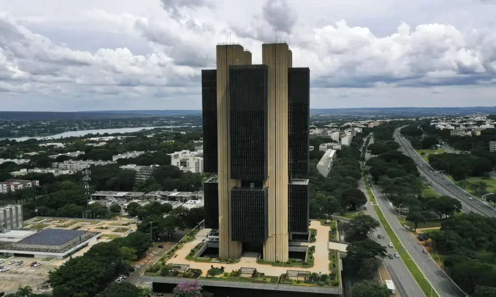 Vista aérea da sede do Banco Central em Brasília, cercada por áreas verdes, vias urbanas e céu com nuvens densas.