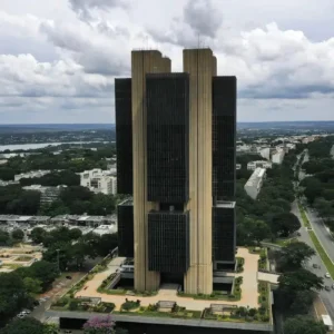 Vista aérea da sede do Banco Central em Brasília, cercada por áreas verdes, vias urbanas e céu com nuvens densas.