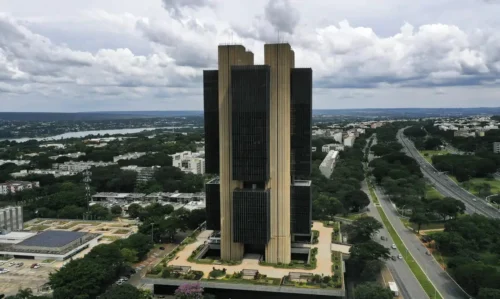 Vista aérea da sede do Banco Central em Brasília, cercada por áreas verdes, vias urbanas e céu com nuvens densas.