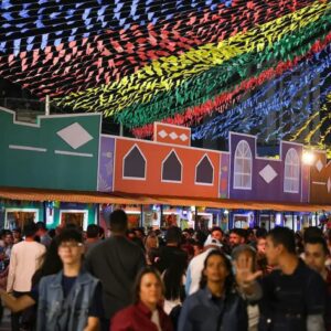 Público participa de festa de São João com bandeirinhas coloridas e decoração junina em espaço de eventos lotado.