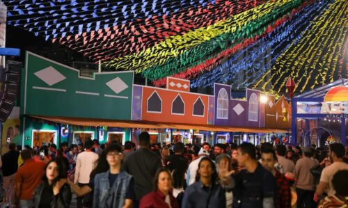 Público participa de festa de São João com bandeirinhas coloridas e decoração junina em espaço de eventos lotado.